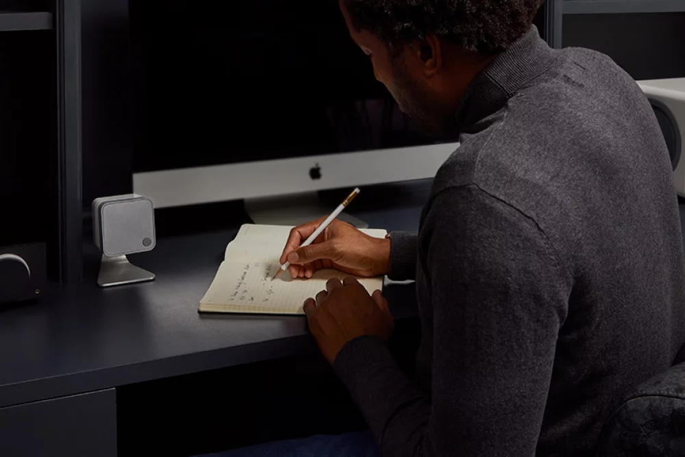man at desk writing in notebook with white minx min 12 speaker on table stand next to him